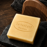 SeifenLust-Bartseife „Savon-de-tête“ auf dunkler Natursteinplatte, mit Holz-Kamm und dunklem Handtuch auf rustikalem Holzuntergrund