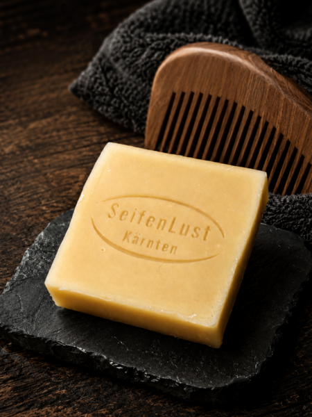 SeifenLust-Bartseife „Savon-de-tête“ auf dunkler Natursteinplatte, mit Holz-Kamm und dunklem Handtuch auf rustikalem Holzuntergrund